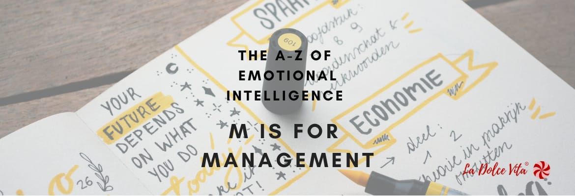 Black text overlaid on a notebook full of business ideas. The words say "The A-Z of Emotional Intelligence. M is for Management"