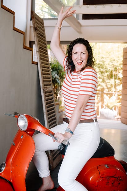 Sarah sits on red Vespa, cheering and smiling